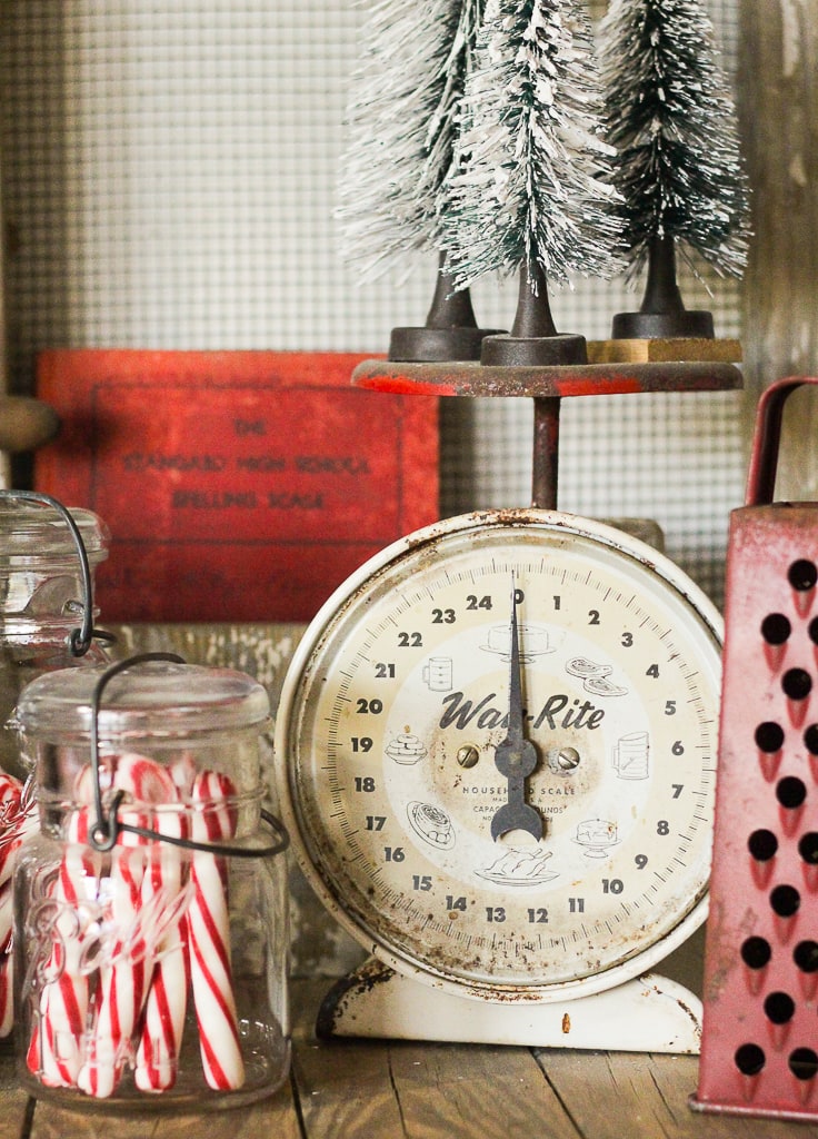 A Christmas vignette an old scale, red grater, old jars with candy canes, a red book and some bottle brush trees.